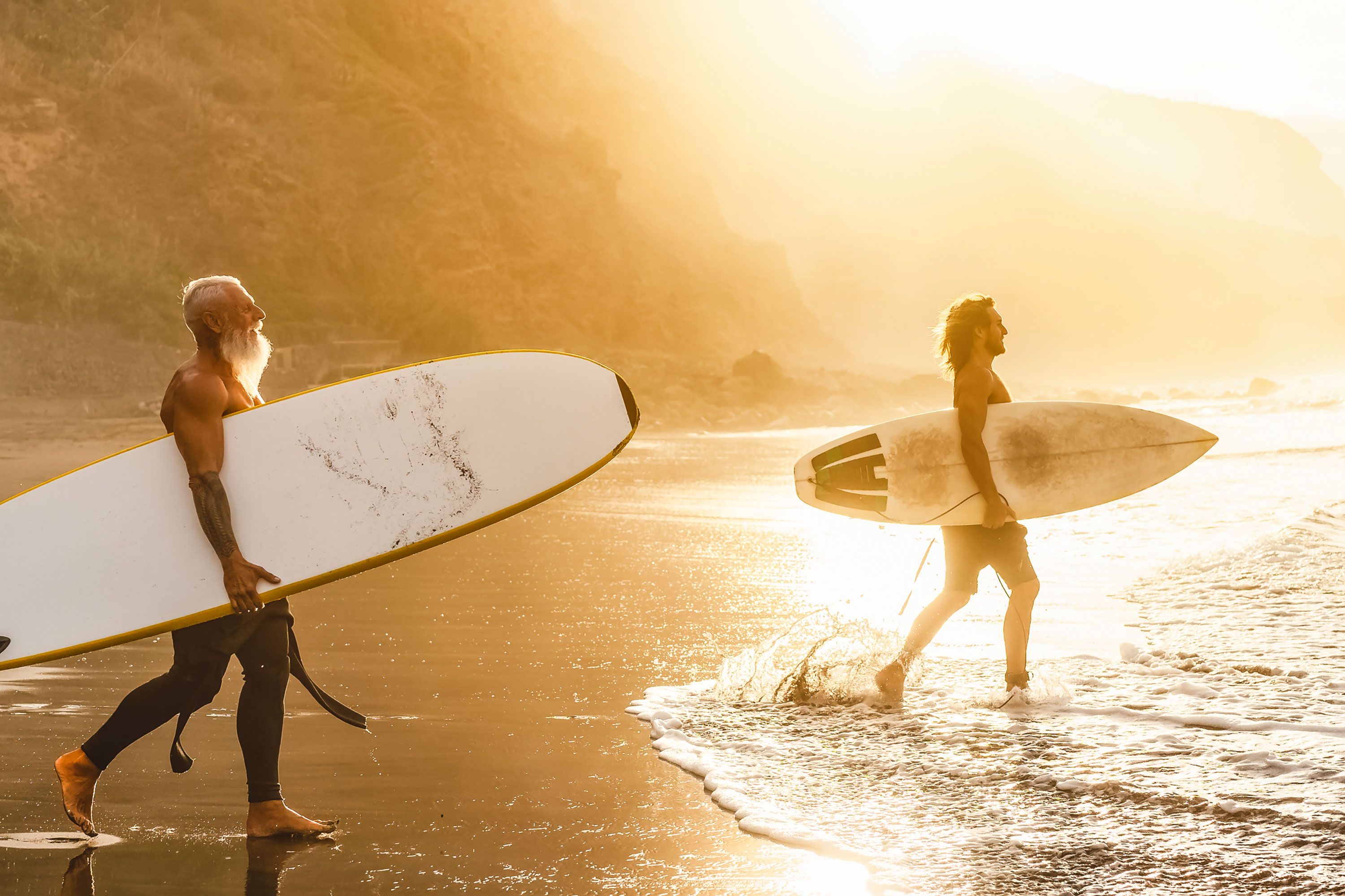 Older man with white beard and man with surfboards head towards the water with the sun shining on them.