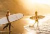 Older man with white beard and man with surfboards head towards the water with the sun shining on them.