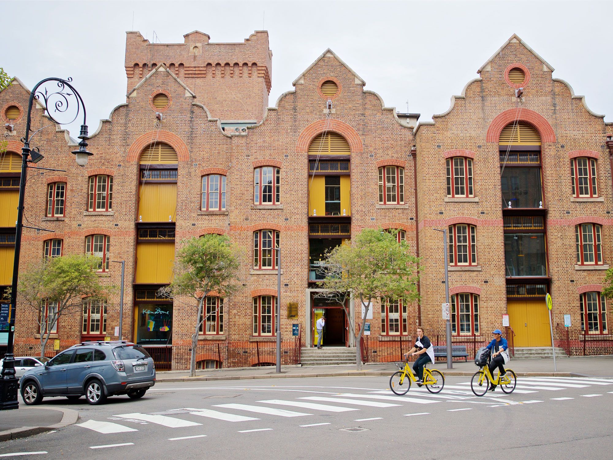 People riding bikes on the road with the brick facade of a building in the background.