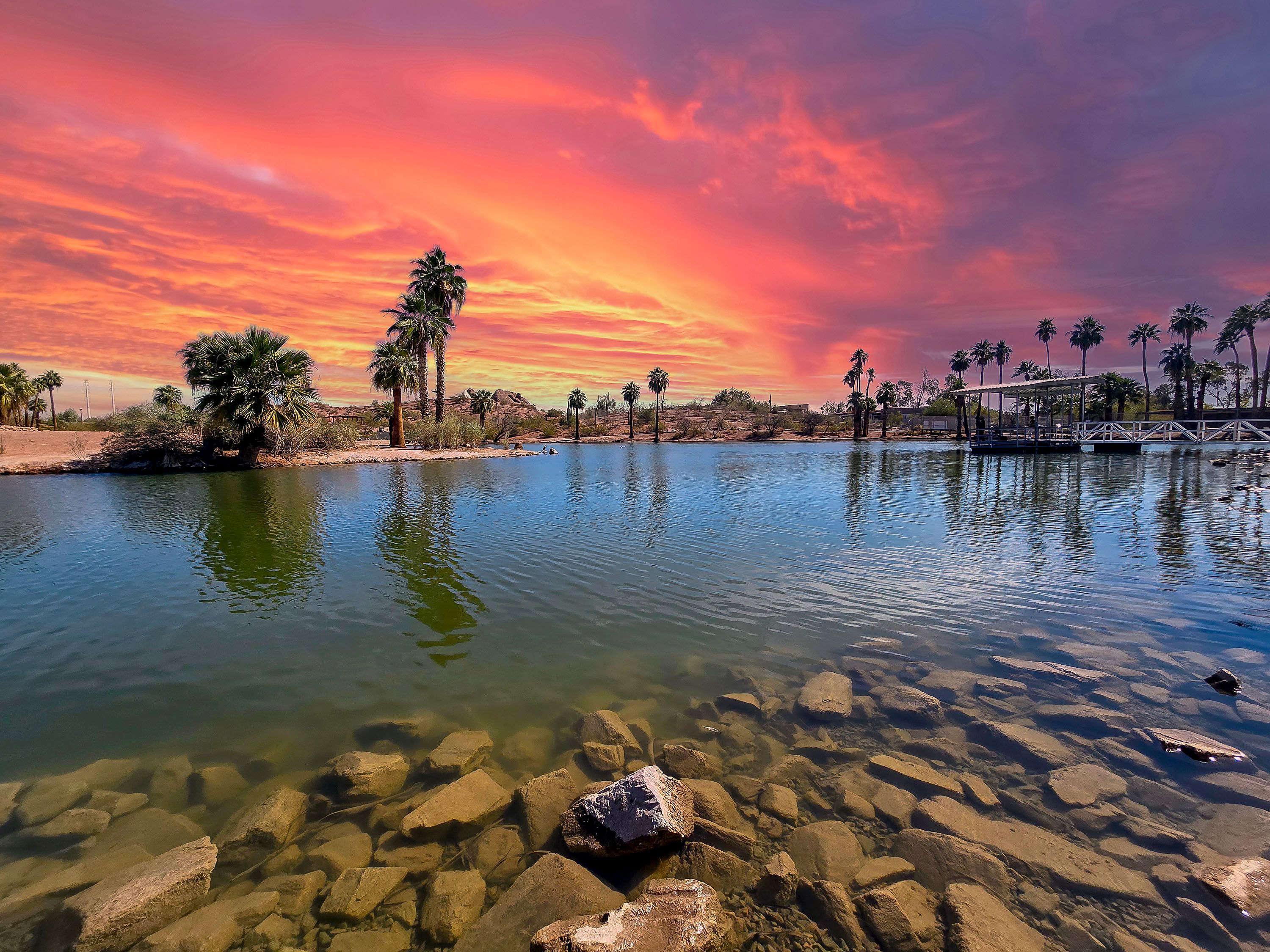Orange and yellow sky with trees and water with rocky shore.