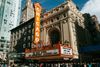 An exterior view of the Chicago Theatre featuring its famous red vertical “Chicago” sign and illuminated marquee on a busy street in downtown Chicago.