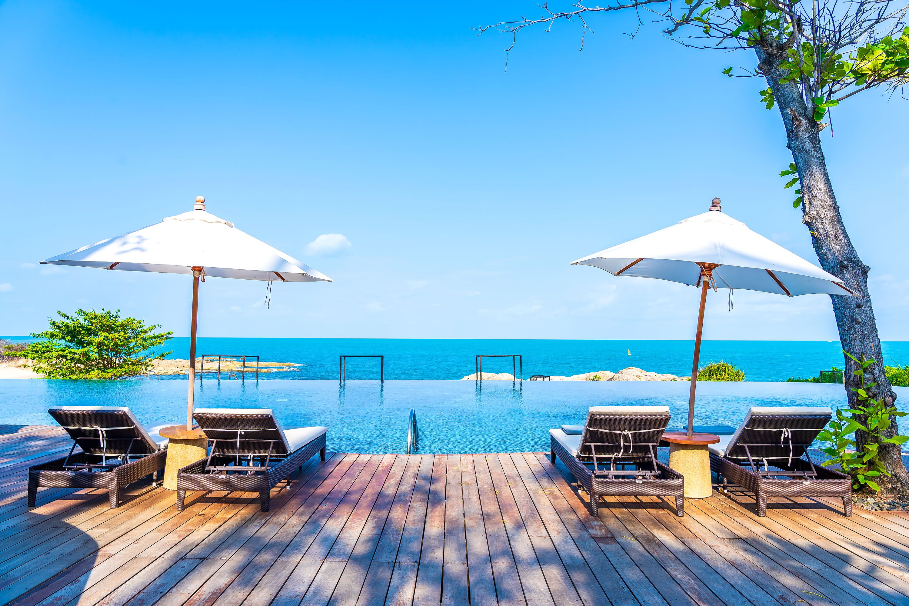 A wooden deck with lounge chairs and umbrellas surrounding an infinity pool overlooking the ocean at a hotel resort.
