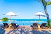 A wooden deck with lounge chairs and umbrellas surrounding an infinity pool overlooking the ocean at a hotel resort.