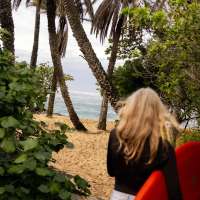 A blonde woman is shown from behind carrying a red surfboard under her arms, through trees leading onto the beach
