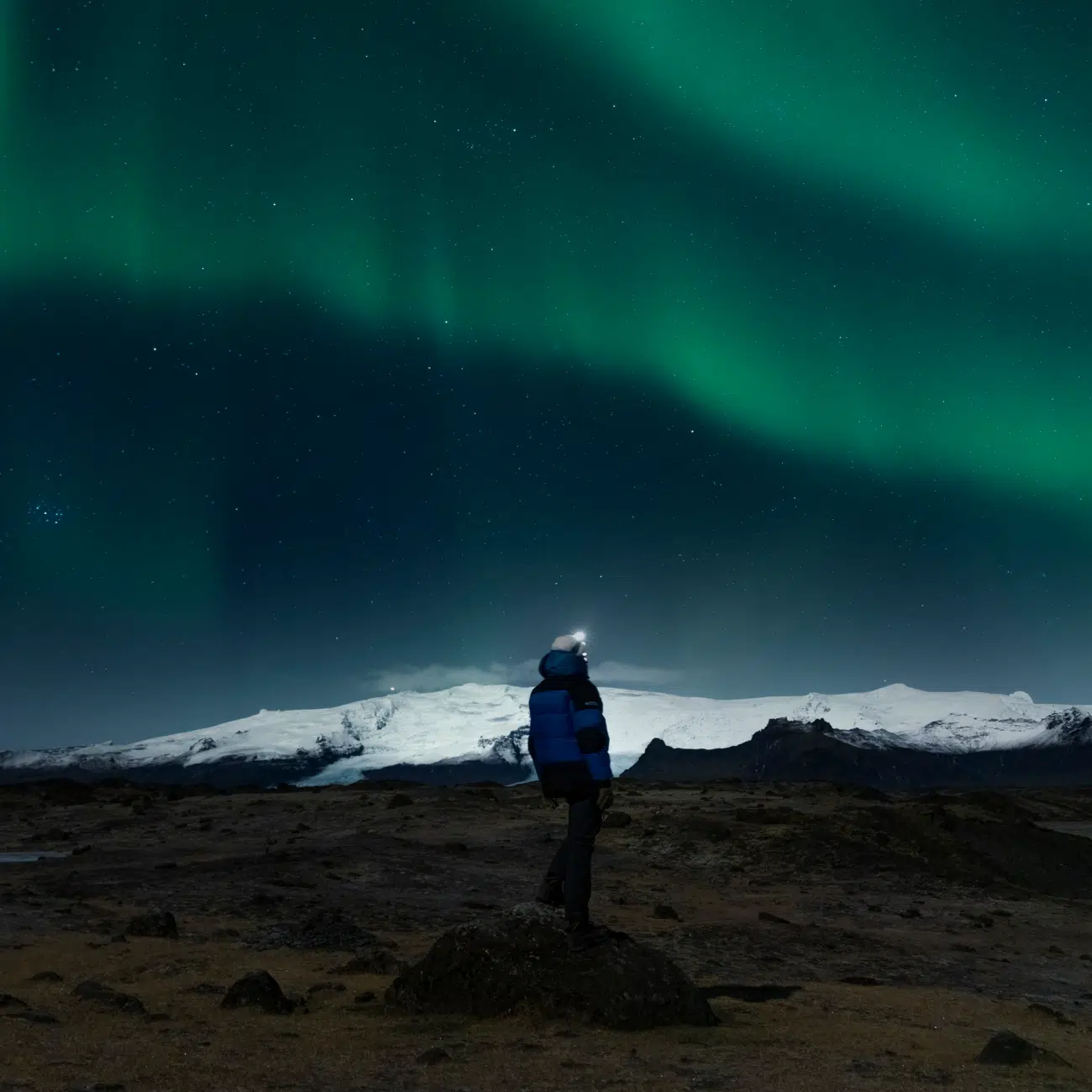 A man stands in a rocky landscape, gazing at the glowing green northern lights