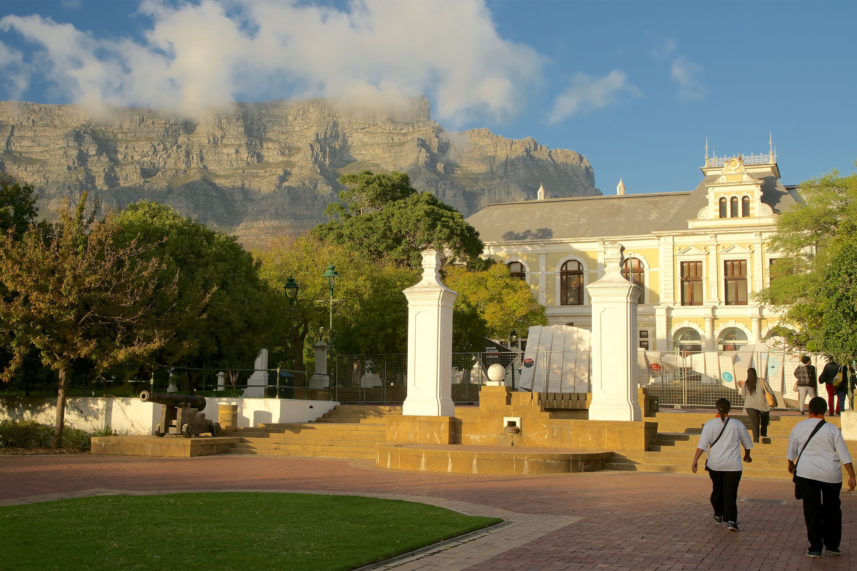 View of the Iziko South African Museum entrance and surrounding trees with people walking in the foreground and Table Mountain towering behind.