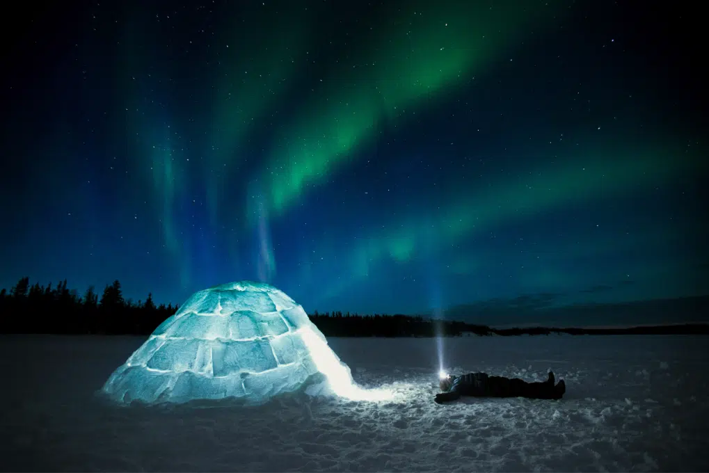 A man lies on the snowy ground admiring the northern lights above, at the entrance to a glowing igloo