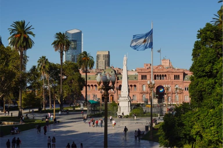 People mull around historic Plaza de Mayo, centered by a white monument and skirted by palms, with a flag flapping in the breeze in the foreground