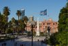 People mull around historic Plaza de Mayo, centered by a white monument and skirted by palms, with a flag flapping in the breeze in the foreground
