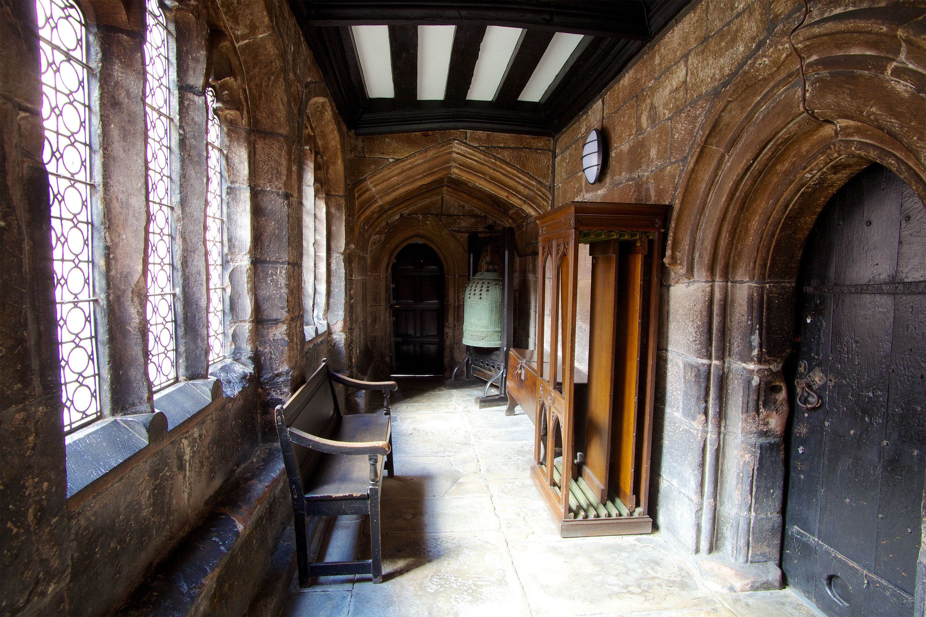 Stone cloister corridor with arched doorways, stained-glass windows, a wooden bench, and an old bell.
