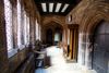 Stone cloister corridor with arched doorways, stained-glass windows, a wooden bench, and an old bell.