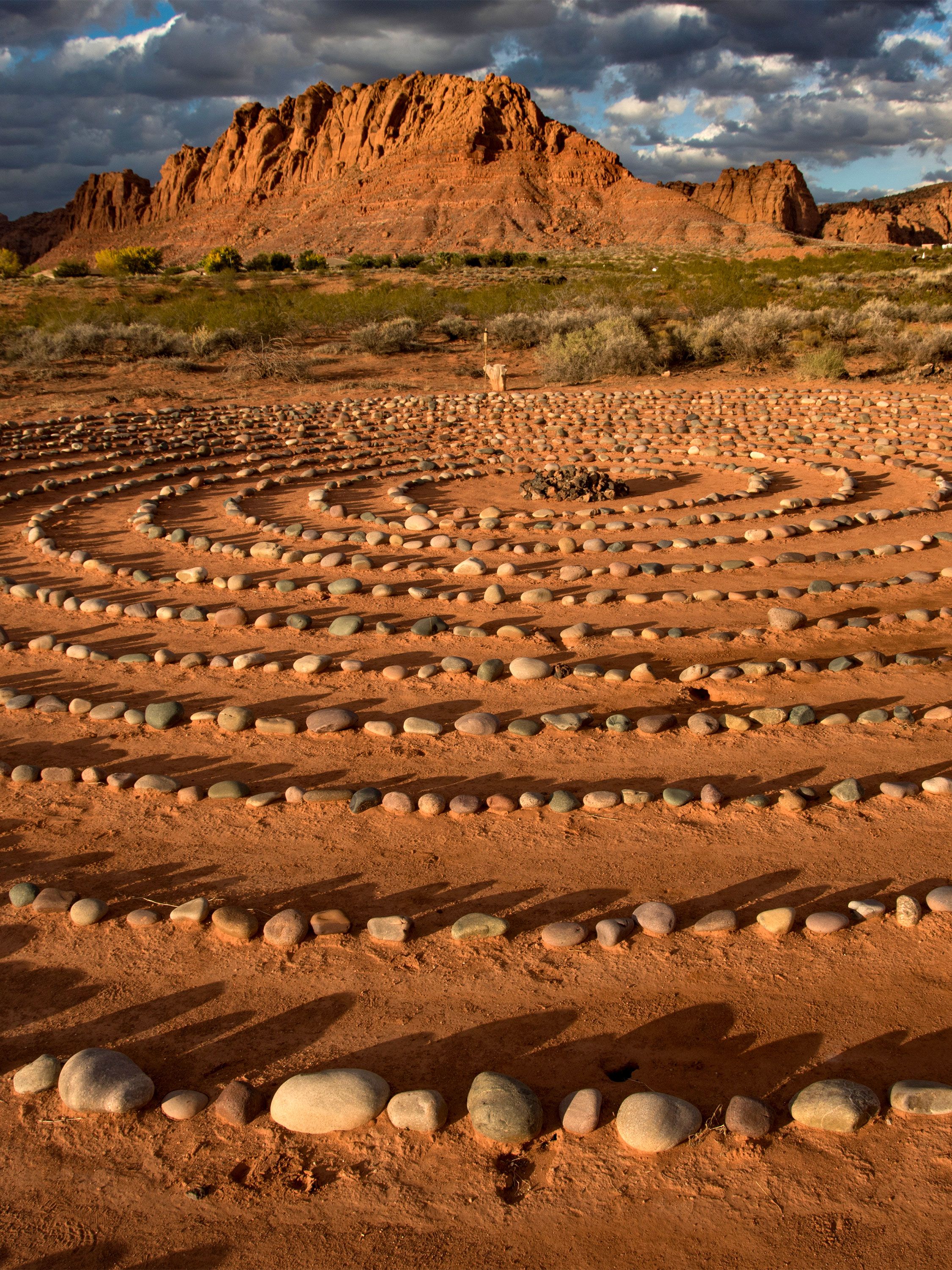 A stone labyrinth spirals across red desert sand beneath rugged sandstone cliffs.