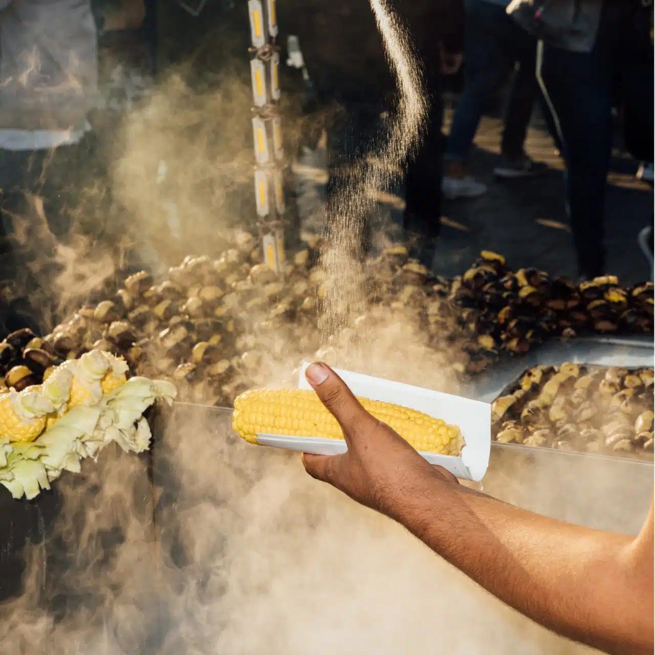 A hand is holding a steaming corn cob in paper, with a market stall selling corn and chestnuts seen behind