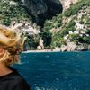 From a boat with a person's blonde hair blowing in the wind on the left, we see the deep blue sea and beach beyond, then the tree-covered cliffs of the Amalfi Coast in Italy