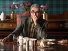 Eugene Levy in a hotel dining room in Scotland, with tartan walls, tartan chair and checked shirt (Image courtesy of Apple TV+)