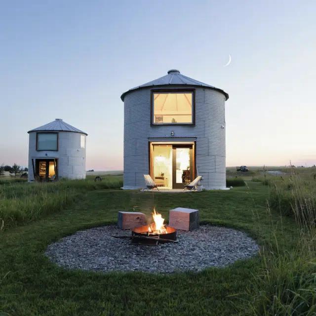 Two converted grain silos in the countryside of Montana at dusk, with a lit campfire burning out front