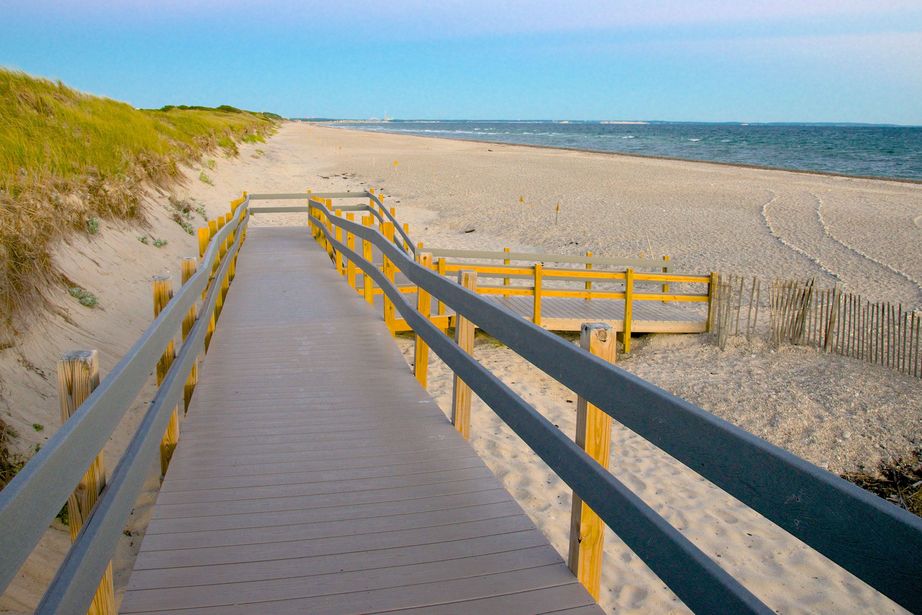 A wooden boardwalk extending over dunes toward a long, quiet beach with gentle waves and grassy sandbanks under a clear blue sky.