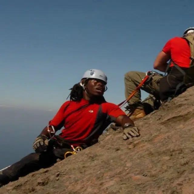 Ade Adepitan in mountaineering gear on a steep slope, with a helper above