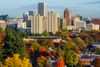 A wide view of Portland, Oregon’s downtown skyline with tall buildings rising above dense fall foliage in shades of red, orange, yellow, and green under a clear, lightly clouded sky.