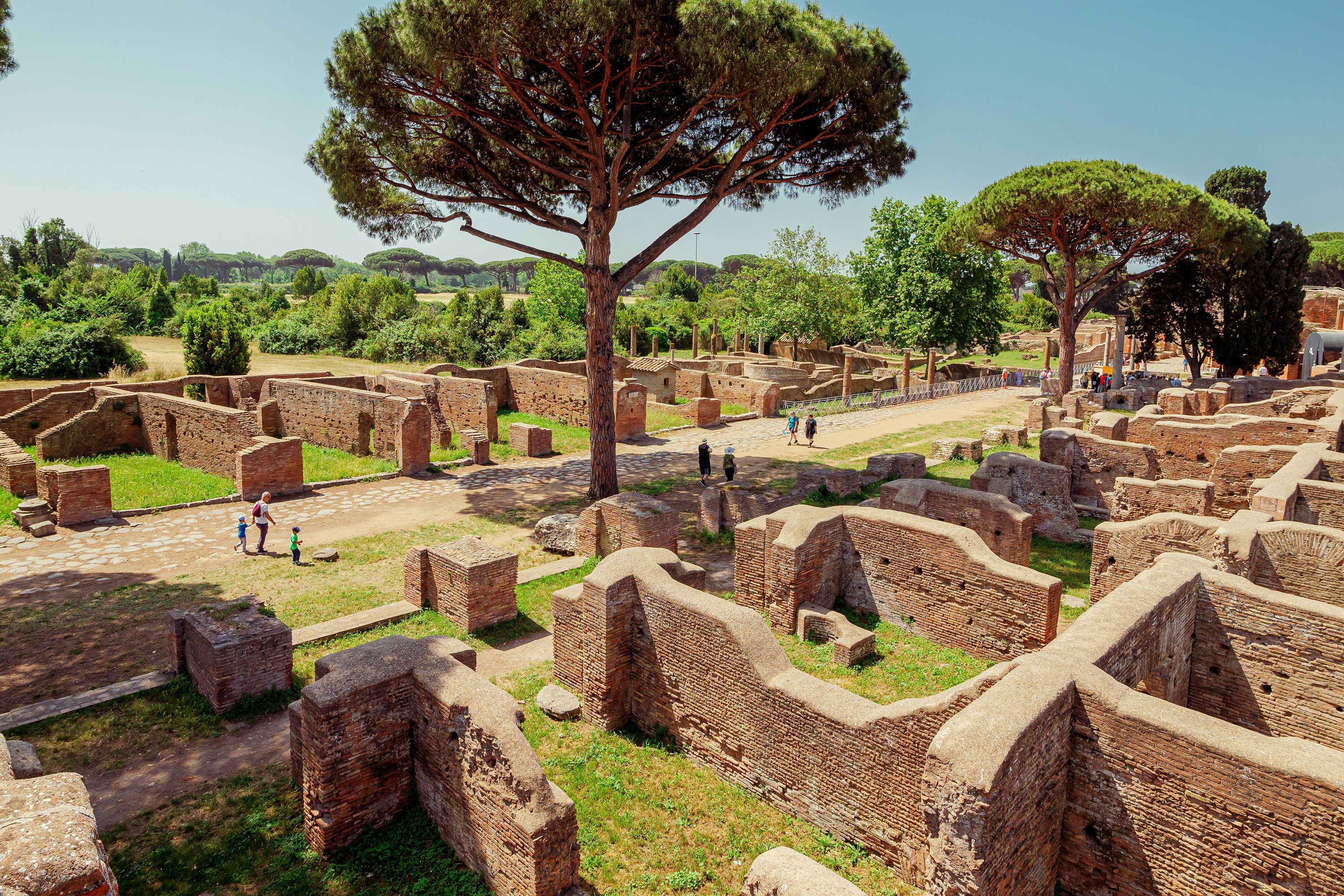People walking down a pathway with old brick walls and a large tree in the center in Ostia Antica, Italy