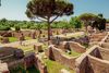 People walking down a pathway with old brick walls and a large tree in the center in Ostia Antica, Italy