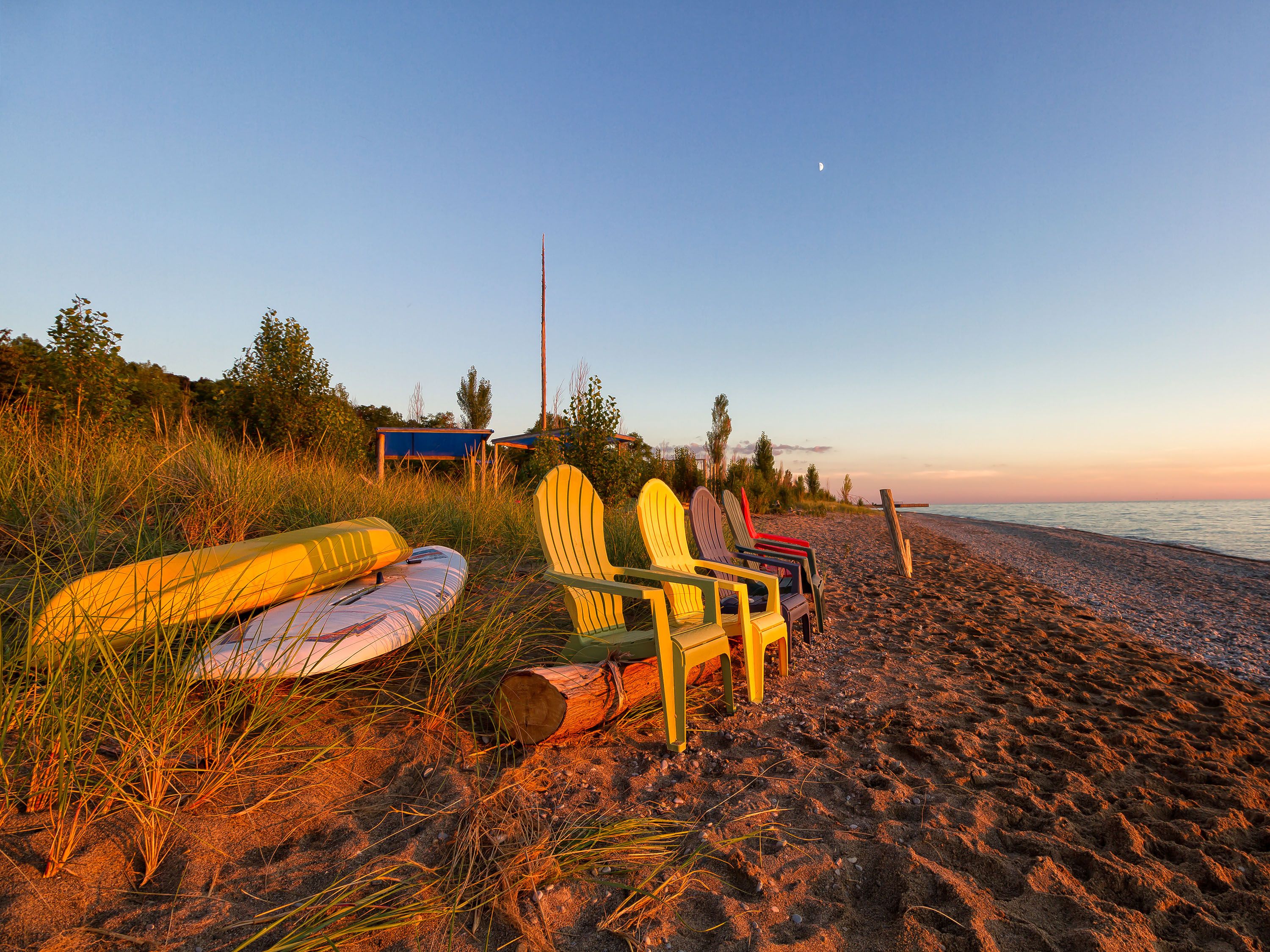 Kayaks and adirondak chairs on the beach as the sun sets.