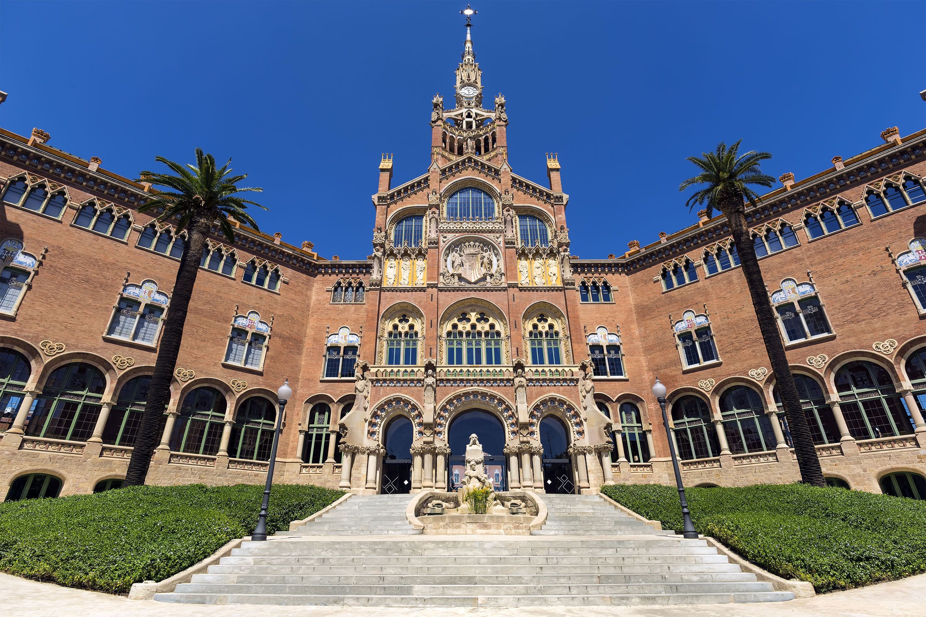 Front view of the Hospital de la Santa Creu i Sant Pau in Barcelona featuring its richly detailed red-brick façade, arched windows, sculptures, and palm trees under a clear blue sky.