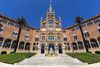 Front view of the Hospital de la Santa Creu i Sant Pau in Barcelona featuring its richly detailed red-brick façade, arched windows, sculptures, and palm trees under a clear blue sky.