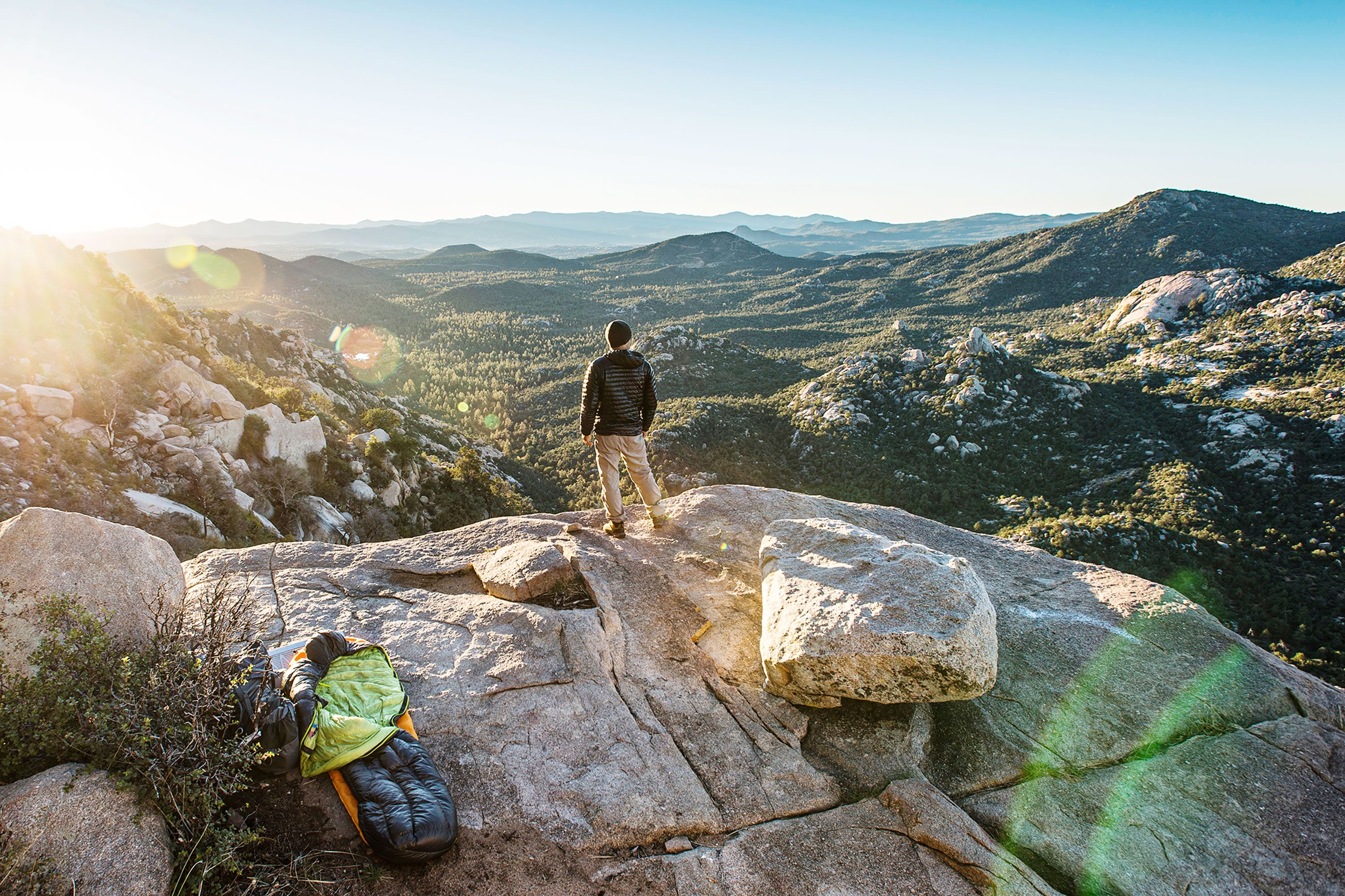 Man with back to camera on rocky outcrop with slepping bag looks out over hills at sunrise.
