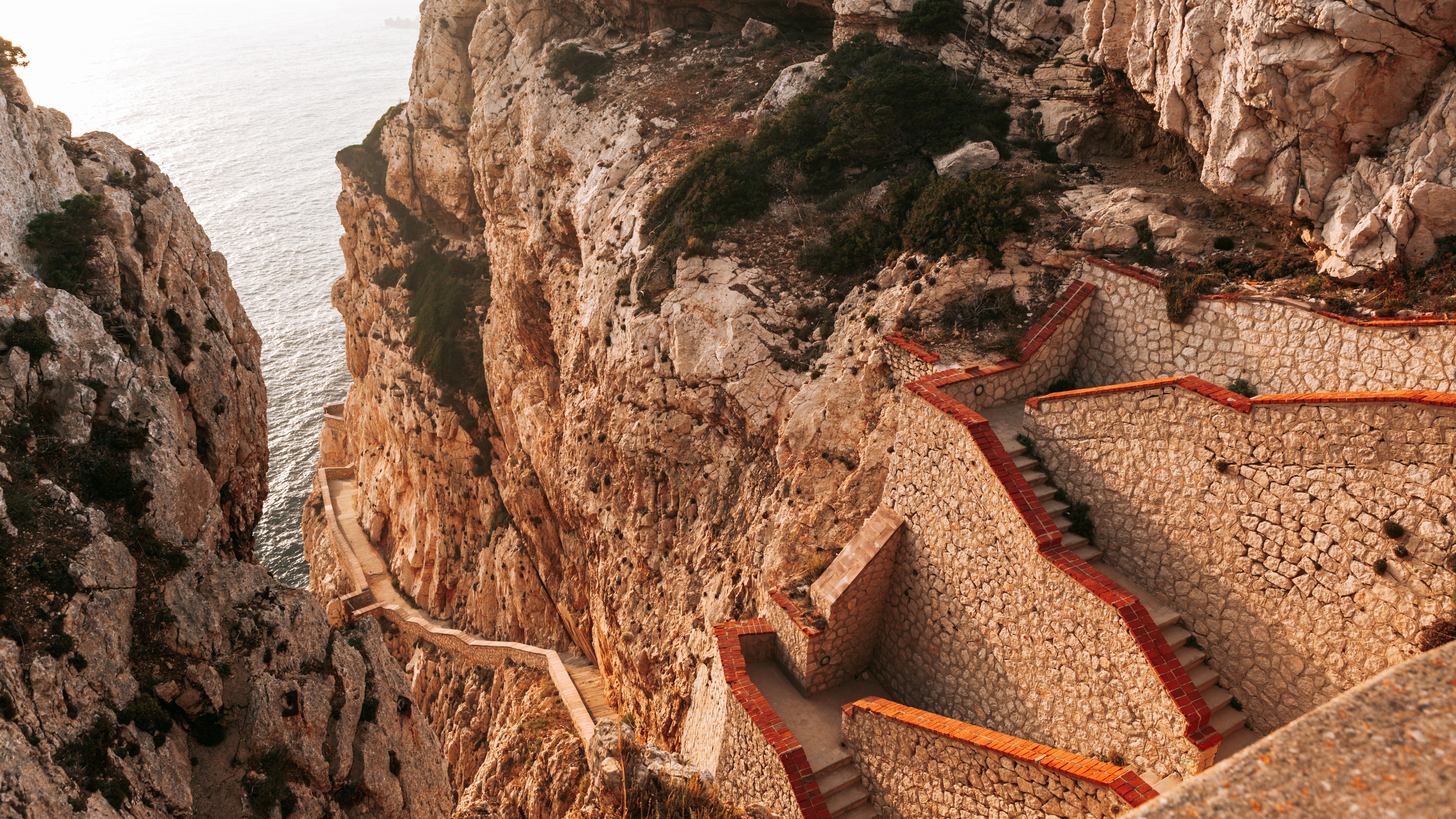 Sardinia, Italy, featuring winding stone paths along rugged cliffs overlooking the sea at sunset.
