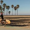 A woman in a long-sleeve swimsuit cycles beside a beach, a wooden surfboard attached to the bike