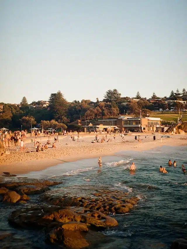 Beneath blue skies on a sunny day, people swim in the sea and others hang out on the golden sands of Bronte Beach in Sydney, Australia