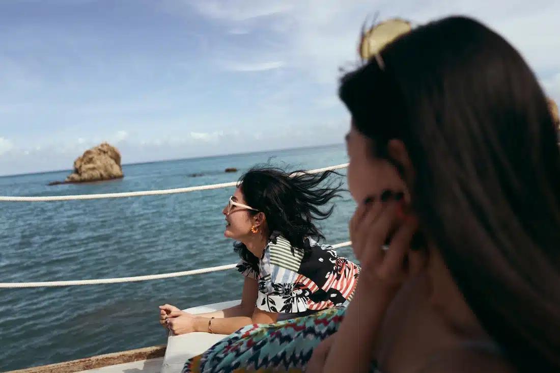 Two women with long dark hair are sailing on a boat over the sea, one seen from behind looking over at the other lying on her front on a sun lounger