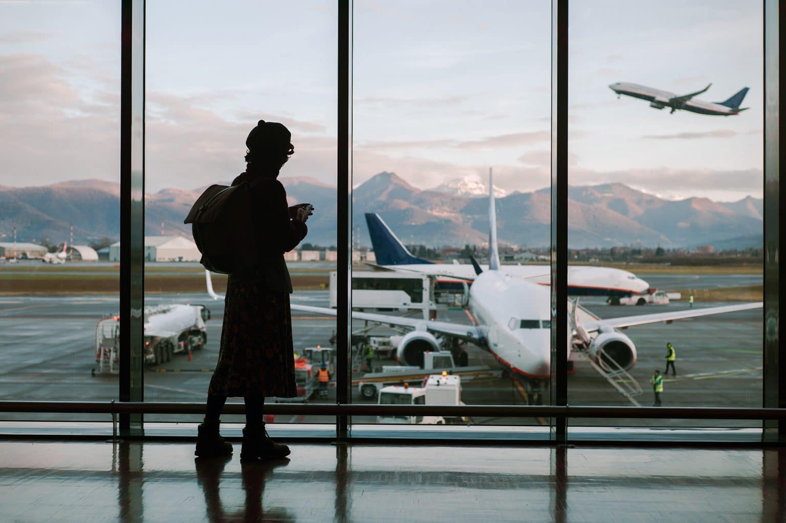 A woman wearing a beret and a backpack looks out of an airport window as a plane takes off