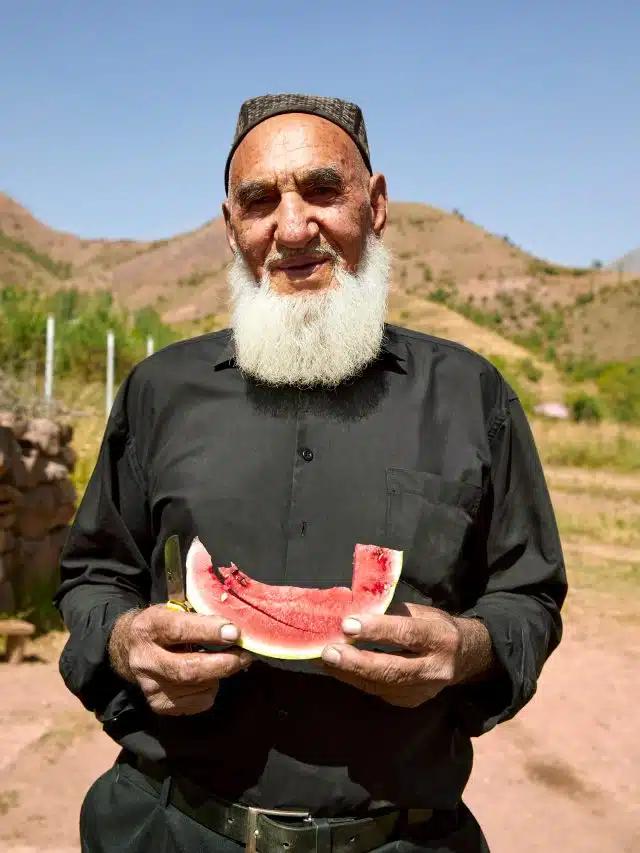 A bearded man wearing black holds a slice of watermelon on a roadside, Tajikistan