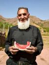 A bearded man wearing black holds a slice of watermelon on a roadside, Tajikistan