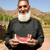 A bearded man wearing black holds a slice of watermelon on a roadside, Tajikistan