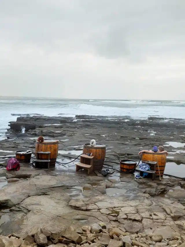 Whisky-barrel seaweed baths sit on the rocky shore in the west of Ireland with an overcast sky