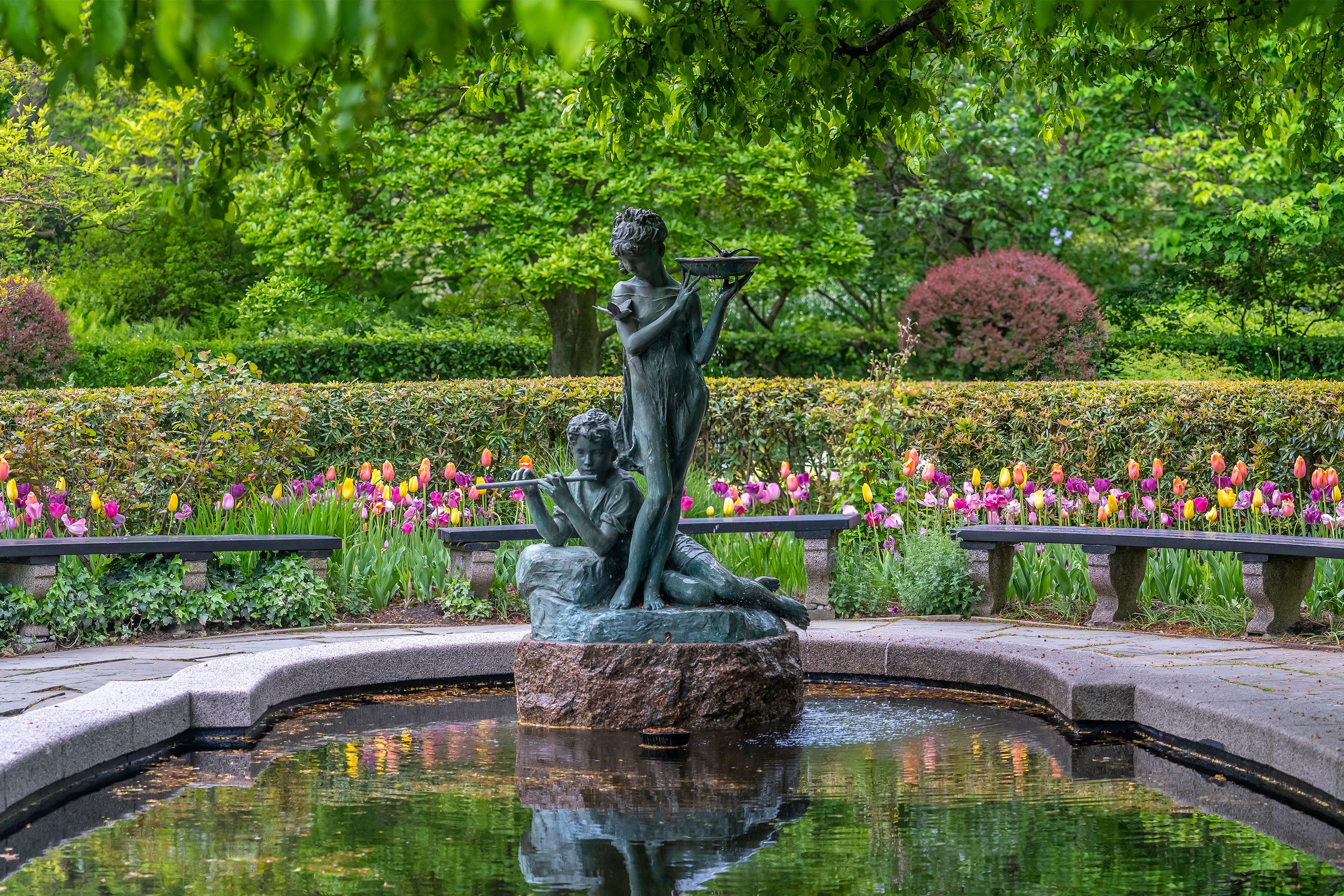 Bronze statue of two children playing instruments at the center of a small reflecting pool, surrounded by colorful tulips and lush greenery in Central Park’s Conservatory Garden.