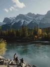 Three people sit on rocks at the edge of the river with fishing equipment as snow-capped Rocky Mountains rise up beyond the opposite bank in Canmore, Canada