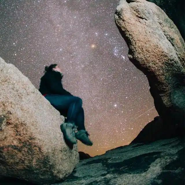 A person sitting on a large rock looks up at a spectacular night sky