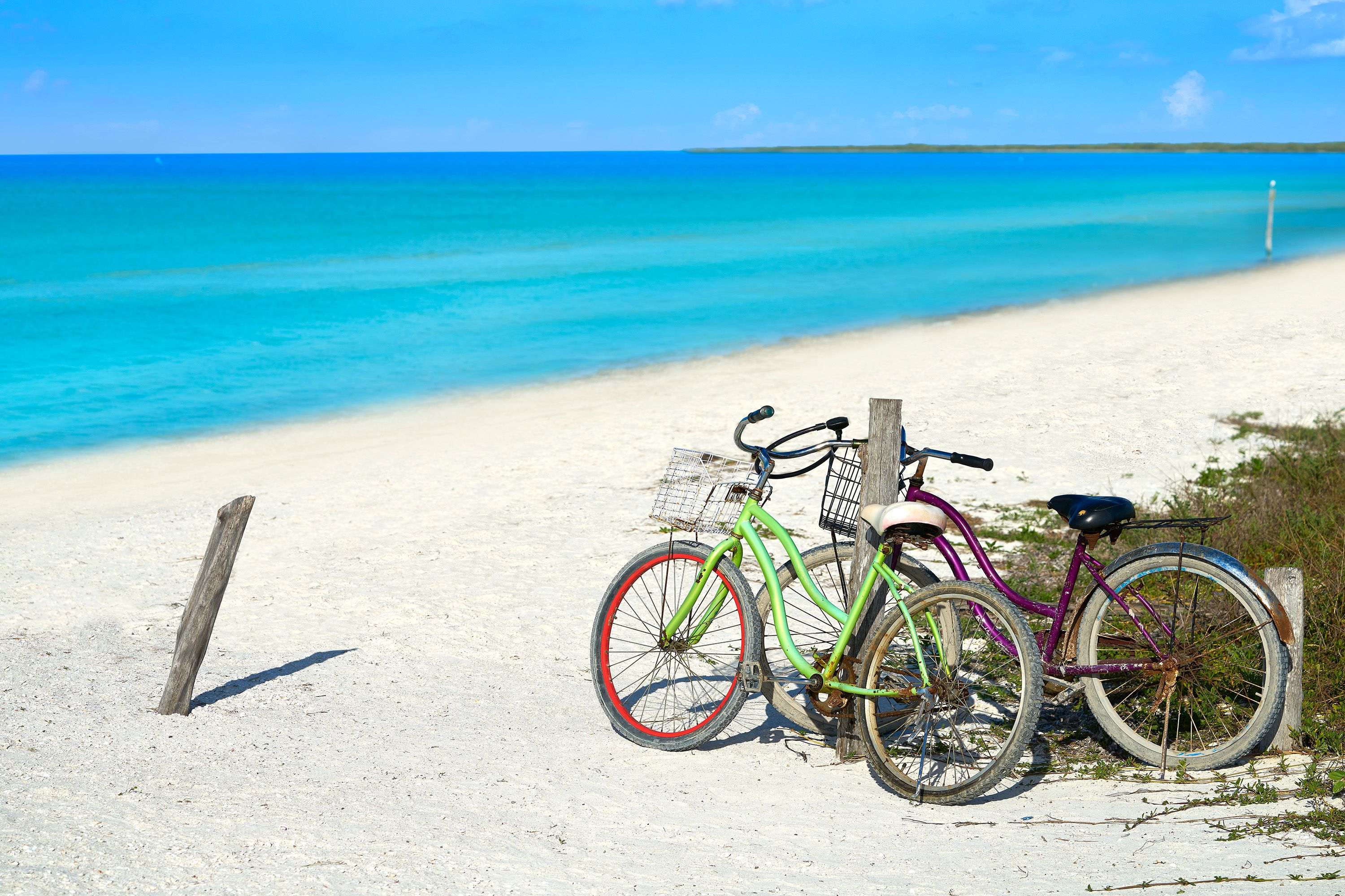 Two bicycles lean against a fence post on a white sand beach with turquoise waters.