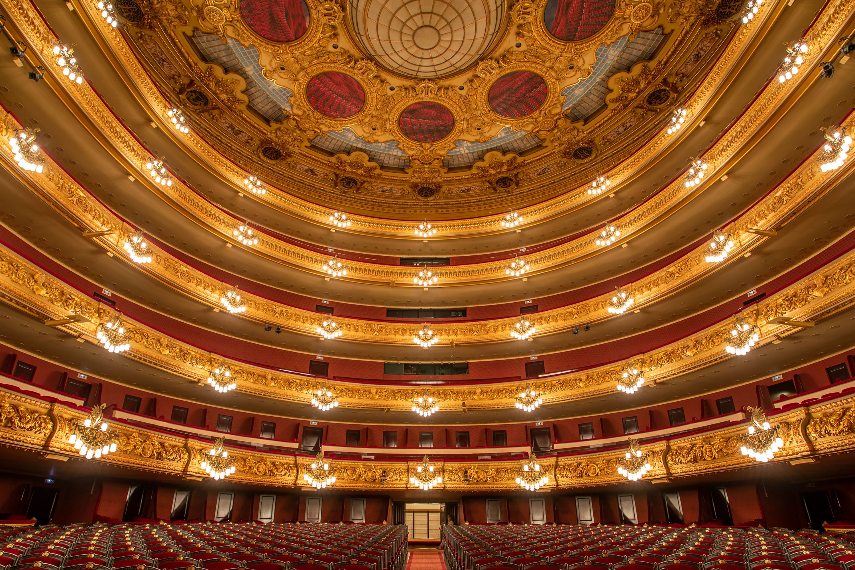 Gilded opera house interior with tiered balconies, chandeliers, and red seats viewed from the stage.