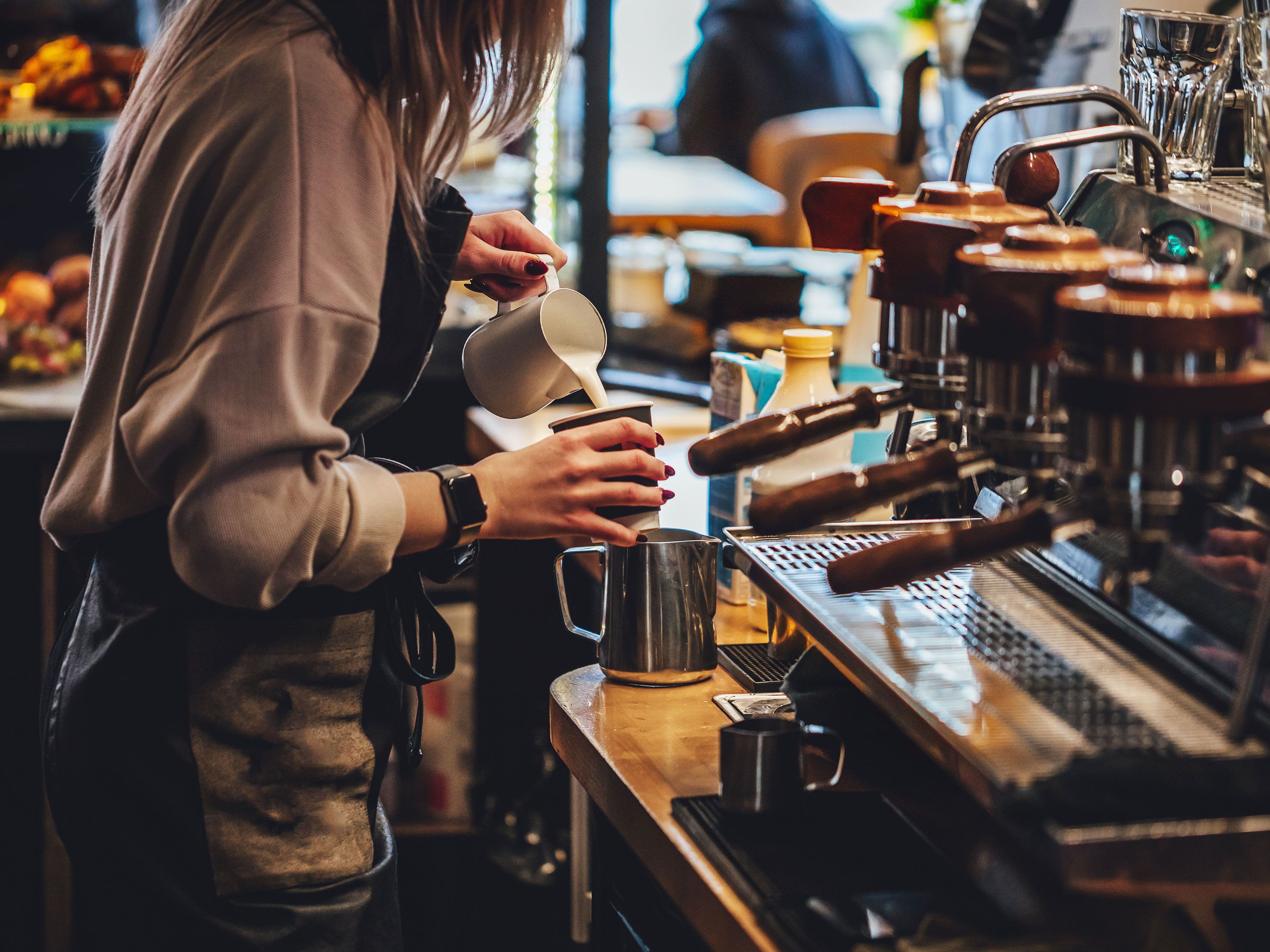 A woman barista pouring milk into a coffee cup next to a large espresso machine.