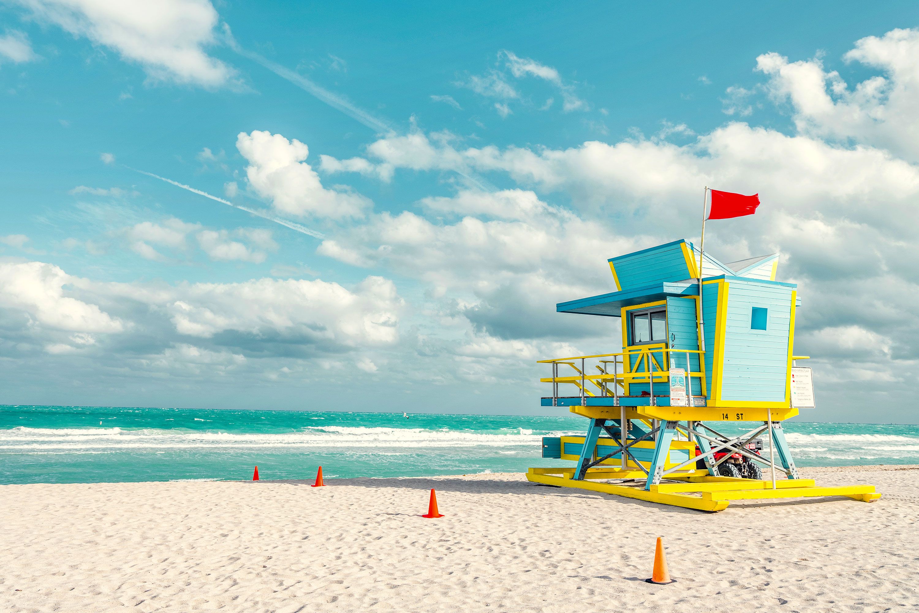 Sandy beach with volleyball net, palm trees looking inland toward neon illuminated hotels under purple sky.