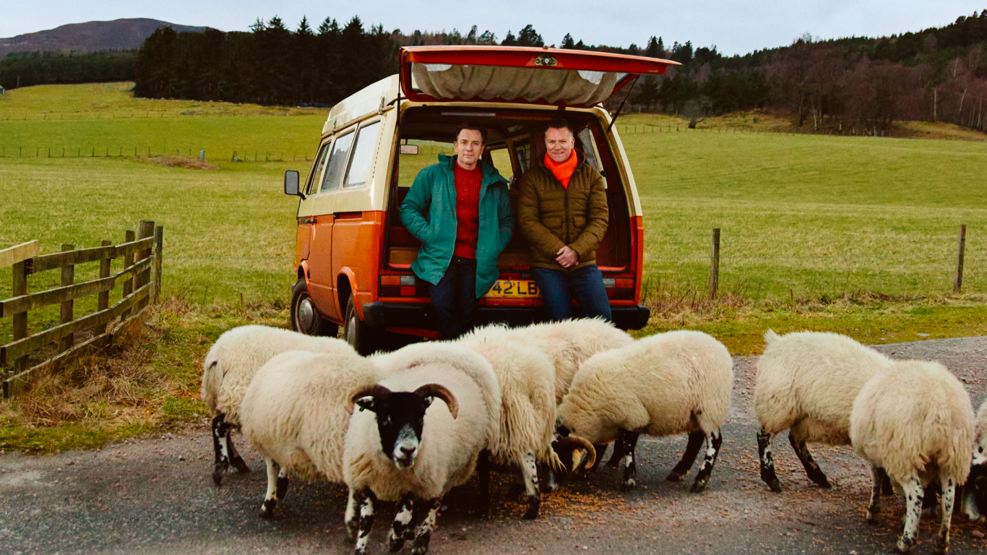 Ewan McGregor and his brother Colin McGregor lean against a orange and cream campervan, a flock of sheep in front of them