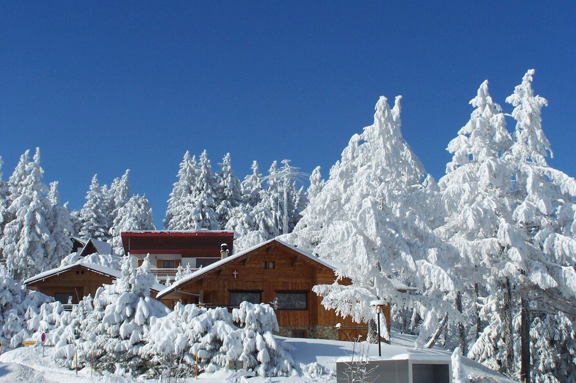Groupe de chalets en bois enneigés au milieu de sapins aux branches givrées en Savoie, où se trouvent de nombreuses locations de vacances Abritel. 