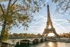 Framed by tree branches in the foreground, a bridge leads toward the Eiffel Tower under a clear blue sky in Paris’s 16th arrondissement