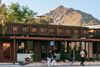 People strolling in front of a wood-paneled building with large square architectural details, lush plants, and Camelback Mountain rising prominently in the background.