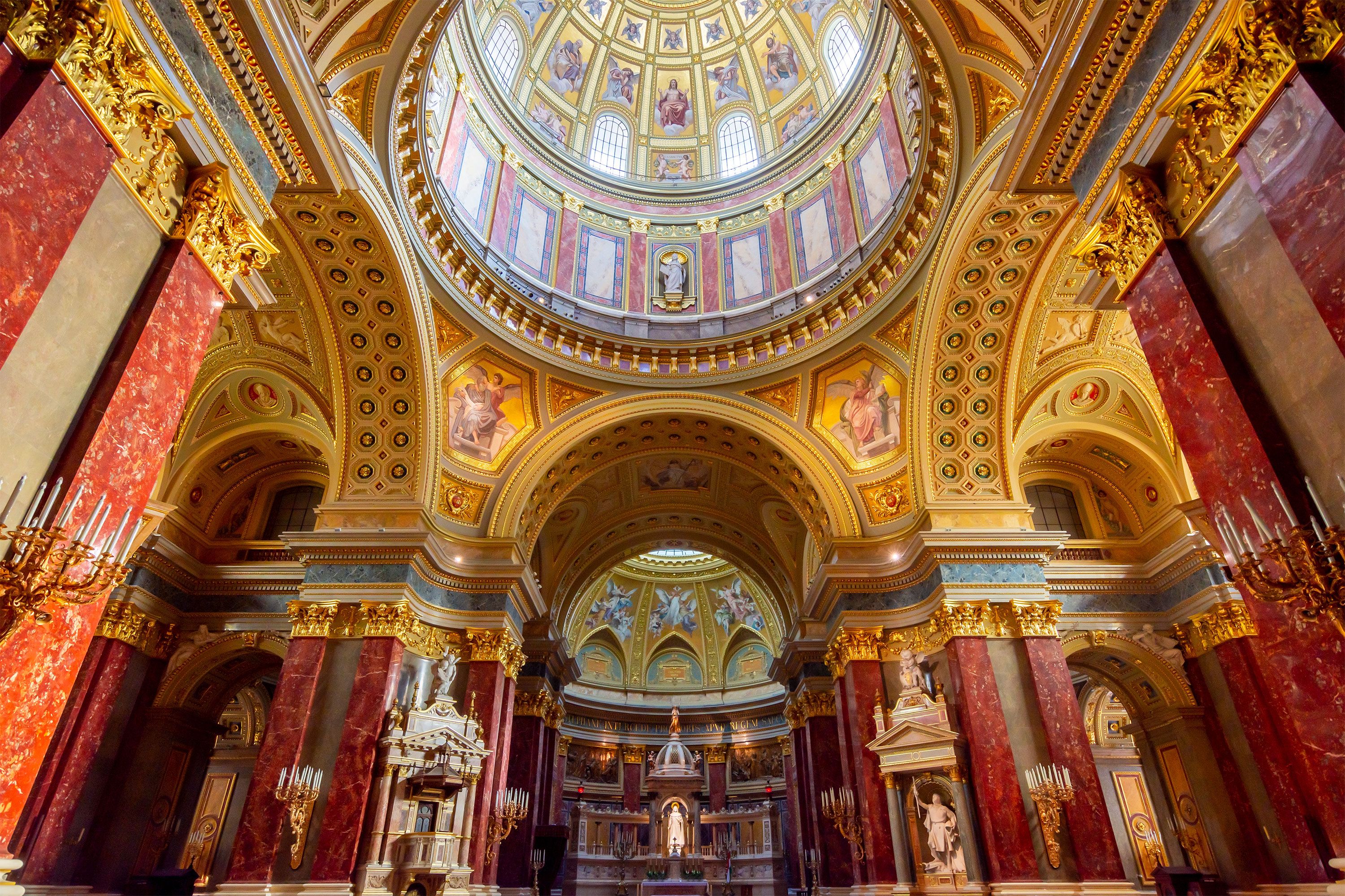 An interior view of St. Stephen’s Basilica in Budapest showing the basilica’s domed ceiling with decorative mosaics, tall marble columns, and richly ornamented arches, bathed in soft natural light.